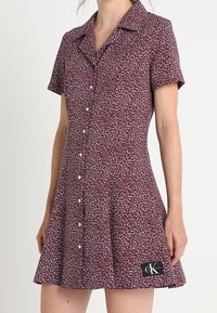 Czarna sukienka w czerwone kwiaty z krótkim rękawem i białymi guzikami z przodu, z naszywką z logo Calvin Klein blisko dołu, noszona przez stojącą osobę.