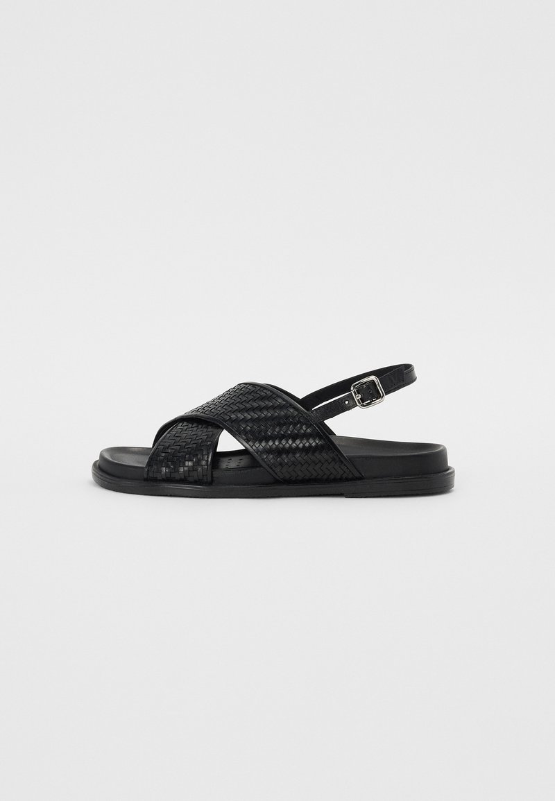 Sorteve svartvevd sandal med kryssbånddesign, justerbar ankelrem og flat gummisåle. Teksturert materiale med glatt finish.