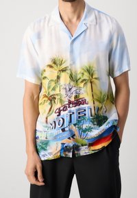 Camisa de manga corta en un tejido ligero que presenta un estampado tropical con palmeras y un cartel de motel, en tonos de azul, amarillo y verde.