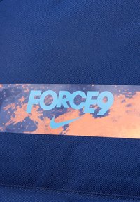 Tumedast kangast, millel on tekstureeritud pind, on horisontaalne silt sinise ja oranži värviga, millele on kirjutatud "FORCE9" ning Nike logo.
