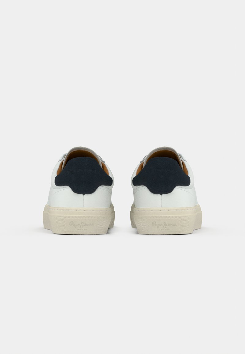 Un par de zapatillas bajas blancas con suelas beige y acentos negros en el talón, mostradas desde la parte posterior sobre un fondo blanco.