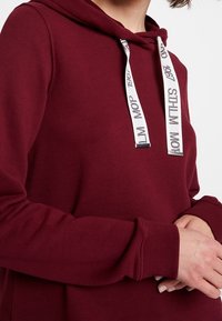 Bordó mikina s klokaní kapsou, kapucí na šňůrky a bílými značkovými akcenty na šňůrkách. Měkká, strukturovaná látka.