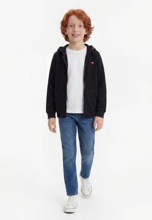 Αγόρι με κόκκινα μαλλιά που περπατάει προς τα εμπρός, φοράει μαύρη ζακέτα με φερμουάρ, λευκό μπλουζάκι, μπλε τζιν και λευκά αθλητικά σε λευκό φόντο.