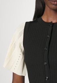 Top smanicato nero a coste con bottoni neri e una manica a sbuffo in crema lavorata a maglia, caratterizzata da motivi a giorno e una texture traspirante.