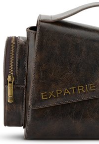 Barna műbőr táska lapos dizájnnal, arany cipzárral és dombornyomott "EXPATRIE" logóval. Varrott részletekkel kiegészítve.