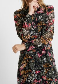 Robe florale en tissu noir transparent, ornée de motifs floraux multicolores en rose, orange et blanc ; détails de col et de poignets boutonnés.