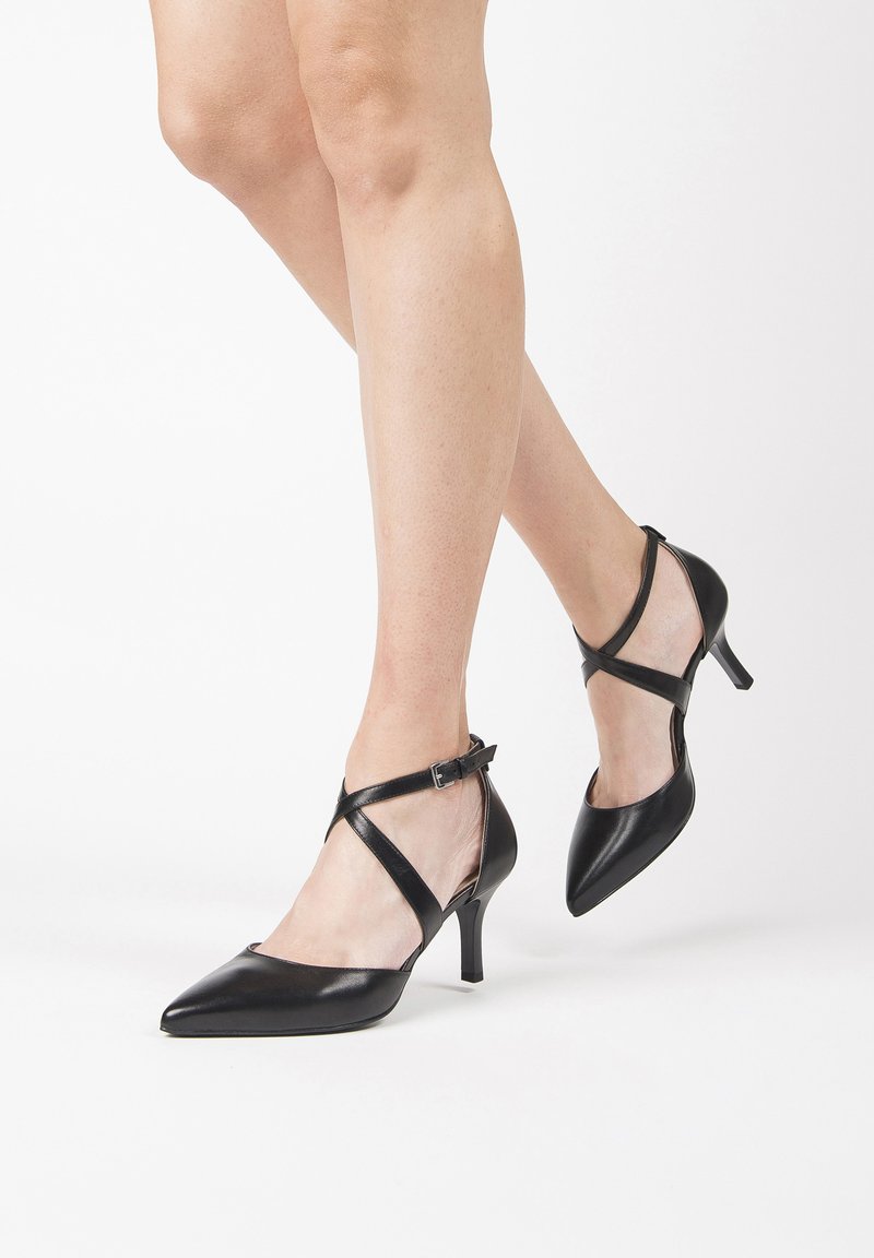 Chaussures à talons hauts en cuir noir avec des pointes pointues, des brides de cheville et un design croisé. Le talon mesure environ 5 cm. Texture lisse et brillante.