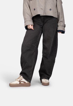 Persoon die zwarte jeans met wijde pijpen, beige en bruine sneakers en een korte geruite jas met opgerolde mouwen draagt, staand tegen een effen achtergrond.