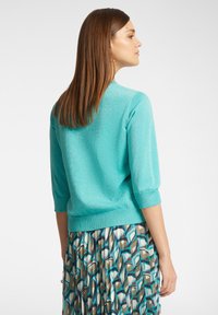 Maglione turchese a tre quarti con una finitura testurizzata e brillante, abbinato a una gonna a pieghe con motivo nei toni di blu e marrone.