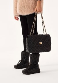 Borsa a tracolla nere trapuntata con hardware dorato, tracolla a catena e forma rettangolare, abbinata a stivali neri trapuntati e pantaloni neri aderenti.