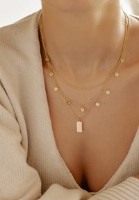 Collares de oro en capas que presentan colgantes circulares planos y texturizados, una etiqueta rectangular, con cadenas delicadas sobre un fondo claro.