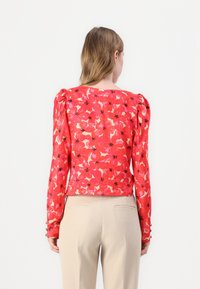 Röd blus med blommönster som har långa ärmar och rynkning på axlarna, i kombination med beige byxor. Tyget verkar lätt och texturerat.