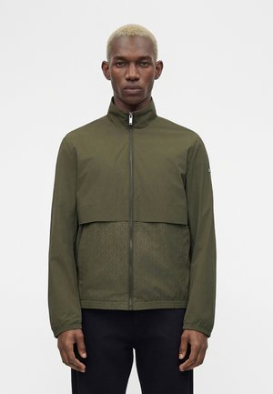 Mężczyzna z krótkimi blond włosami, ubrany w oliwkową kurtkę na zamek z wysokim kołnierzem i czarne spodnie, stojący na białym tle.