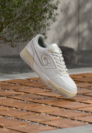 Biały skórzany sneaker o niskim profilu, z perforowanym noskiem, jasnobeżowymi akcentami i teksturowaną gumową podeszwą, przedstawiony na tle drewnianej powierzchni.