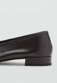 Zapato bajo de cuero negro con tacón bajo, punta redondeada y un simple detalle de costura, exhibido sobre un fondo gris claro y liso.