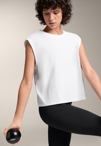 Weißes ärmelloses Oberteil mit lockerer Passform, kombiniert mit schwarzen Leggings. Das Modell hält ein schwarzes Fitnesszubehör in der rechten Hand.