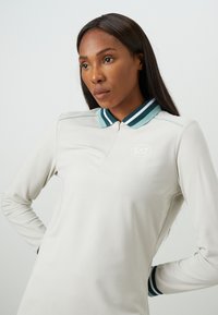 Vrouw met lang, donker haar die een lichtbeige polo met lange mouwen draagt, voorzien van een groene gestreepte kraag en het EA7 Emporio Armani-logo.