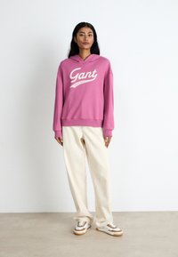 Pink hoodie with white "Gant" logo, loose fit, paired with cream-colored wide-leg pants and white sneakers with brown accents.