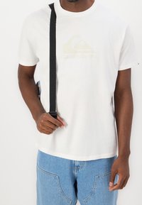 Man som bär en vit Quiksilver-t-shirt och ljusblå jeans, håller en svart rem över axeln mot en enkel bakgrund.