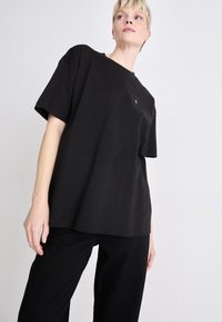 Svart oversized t-skjorte i bomullsblanding med korte ermer, rund hals og liten logodetalj på brystet, sammen med svarte vide bukser.