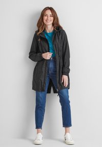 Black waterproof jacket with a hood, featuring a subtle geometric pattern, adjustable waist, and front pockets. Paired with a blue top and jeans.