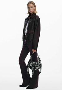 Czarna skórzana kurtka z kołnierzem, zestawiona z głęboko burgundowymi dżinsami w fasonie dzwonów. Trzyma torebkę w czarno-białą fakturę z unikalnym wzorem paska.