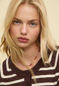 Modelka s blond vlasy a modrýma očima, oblékající hnědo-bílý pruhovaný cardigan. Záběr zblízka, který zdůrazňuje texturu pleti a šperky.