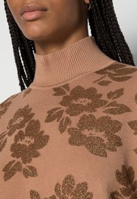 Pull rose clair avec un col montant côtelé, présentant des motifs floraux bronze foncé et un tissu texturé. Gros plan sur le haut du vêtement.