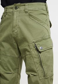 Olive green cargo pants with multiple pockets, including a flap pocket with snap buttons and a vertical zipper pocket on the thigh.
