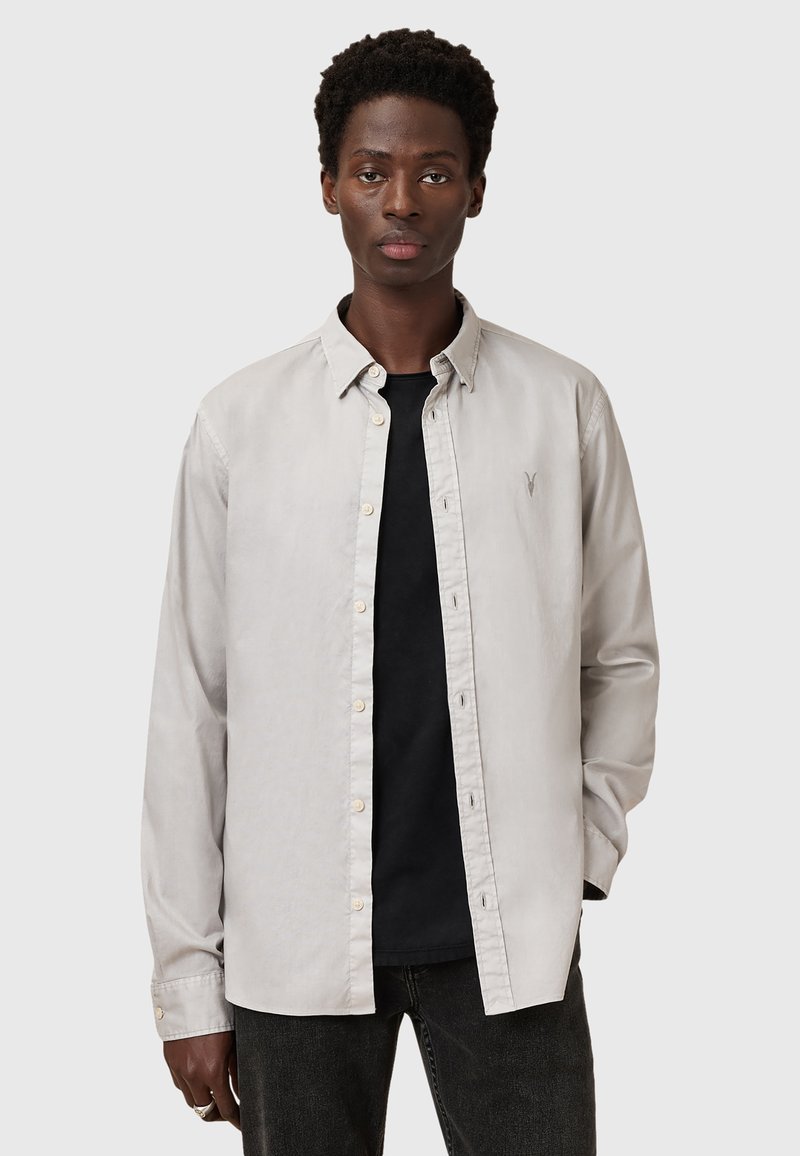 Lichtgrijs button-up overhemd met een kraag, voorzien van een klein logo op de borst, gedragen met een zwart t-shirt eronder en donkere jeans.