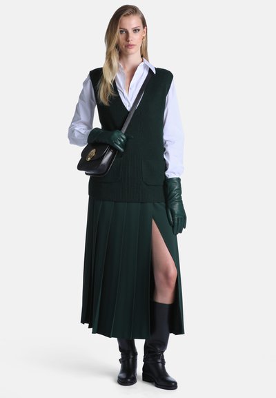 Femme portant une jupe plissée vert foncé avec une fente, un gilet en maille assorti, une chemise blanche, des gants verts, des bottes noires et un sac à épaule noir sur fond blanc.