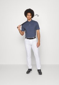 Marineblå polo-skjorte med korte ermer, hvite skreddersydde bukser, svart belte og svarte golfsko. Holder et golfkølle over skulderen.