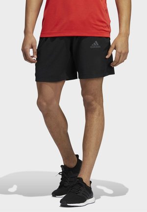 Homme portant un short noir Adidas, des chaussures de sport noires et un t-shirt rouge, debout sur un fond blanc.
