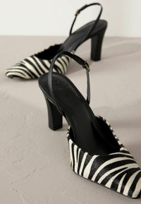 Musta-valkoinen raidallinen slingback-korkokenkä, jossa on teräväkärkinen muotoilu ja paksu korko. Teksturoitu kangas, joka on varustettu kiiltävällä mustalla hihnalla ja solkikoristeella.