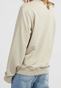 Personne portant un sweat-shirt beige ample associé à un jean en denim bleu clair, vue de dos sur un fond uni.