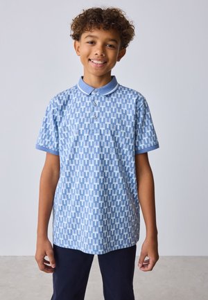 Lächelnder Junge mit lockigem Haar, der ein hellblaues gemustertes Poloshirt und dunkle Hosen trägt, steht vor einem einfarbigen hellen Hintergrund.