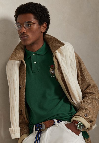 Zelena polo majica z vezenim logotipom, podložena pod bež in rjavim plaščem z ribjo kostjo, ki ima pletene poudarke in modni pas.