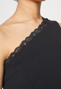 Černý top na jedno rameno s jemným černým krajkovým lemem podél výstřihu. Hladký materiál s přiléhavým střihem a mírným drapováním.