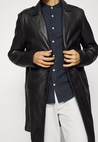 Cappotto di pelle nera con collo a punte, due bottoni e spalline sulle spalle. Indossato sopra una camicia a bottoni blu scuro e pantaloni bianchi.