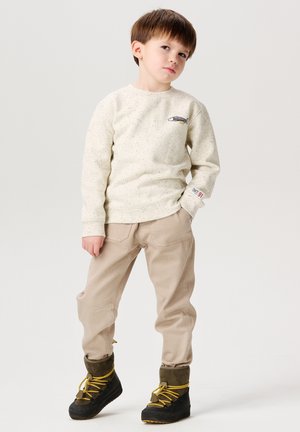 Felpa cremoso macchiata con design di pesce, pantaloni beige e stivali neri con lacci gialli. Bambino in posa con le mani sui fianchi.