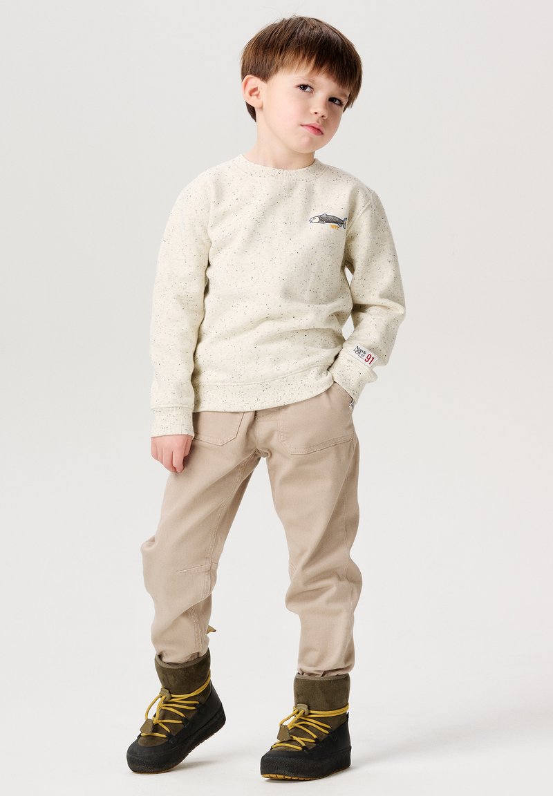 Cremefarbenes Sweatshirt mit Fischdesign, beige Hose und schwarze Stiefel mit gelben Schnürsenkeln. Kind posiert mit den Händen auf den Hüften.