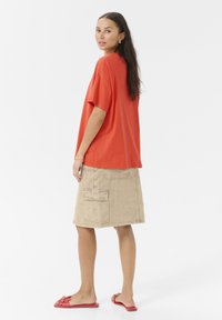 Rød oversized t-shirt parret med en beige cargo-nederdel med to sidelommer. Modellen har røde slide-sandaler på med en dekorativ detalje.