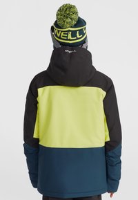 Veste extérieure colorée avec des panneaux noirs, jaune vif et bleu marine, dotée d'une capuche et d'un logo au dos. Bonnet en tricot avec pompon sur la tête.