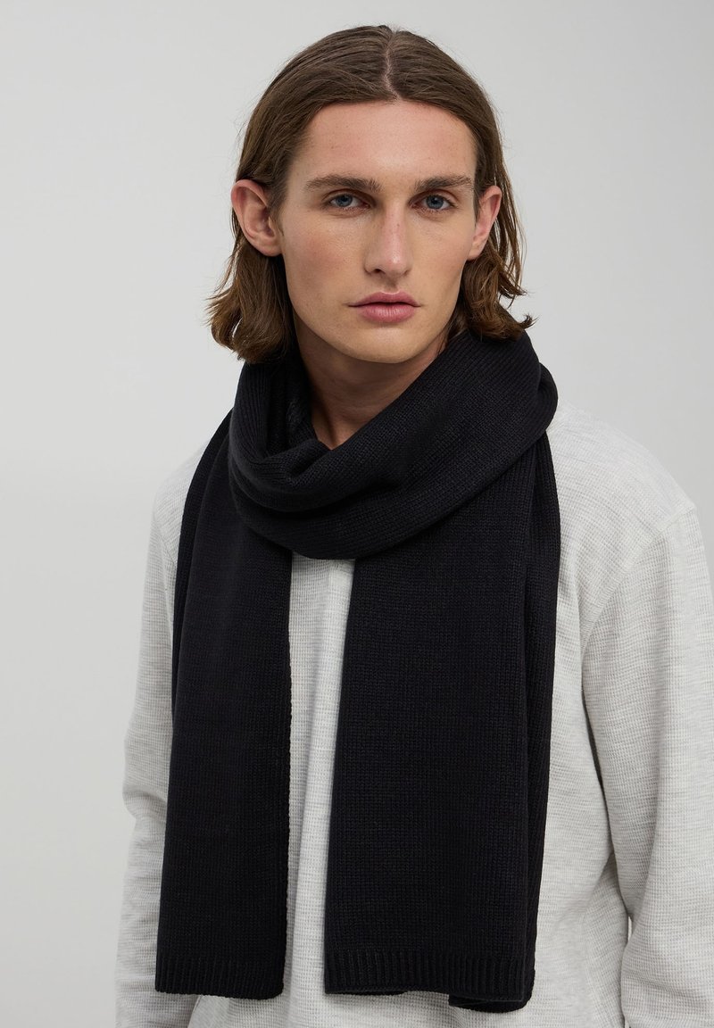 Sciarpa nera lavorata a maglia con una texture grossa, forma ampia e bordi a coste, drappeggiata intorno al collo di una modella che indossa un maglione di colore chiaro.