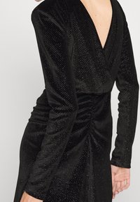 Robe en velours noir avec des manches longues, un dos en V profond, et des fronces subtiles à la taille, ornée d'accents scintillants.