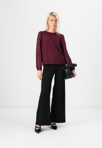 Burgundy textured long-sleeve top with leaf pattern, paired with black wide-leg pants and pointed black flats. Holds a small black handbag.