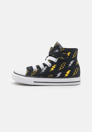 Zapatilla alta negra con un patrón de rayos amarillos y grises, tapa de punta de goma blanca y cordones blancos. Correa de velcro en el cuello.