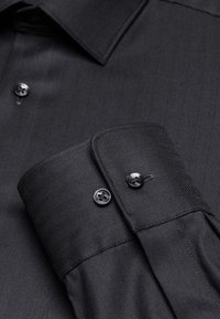 Camicia nera con motivo a spina di pesce, colletto button-down e polsini con due bottoni. Tessuto liscio con una leggera lucentezza.