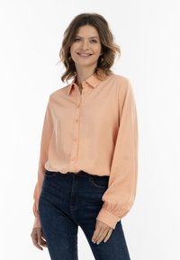 Peachfarvet bluse med krave, knaplukning foran og lange ærmer med samlede manchetter, sammen med mørkeblå jeans. Glat stoftekstur.