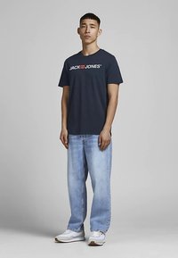 Μπλε βαμβακερό μπλουζάκι με το λογότυπο "JACK & JONES" σε λευκό και κόκκινο, συνδυασμένο με τζιν παντελόνι ανοιχτού μπλε και λευκά αθλητικά παπούτσια.